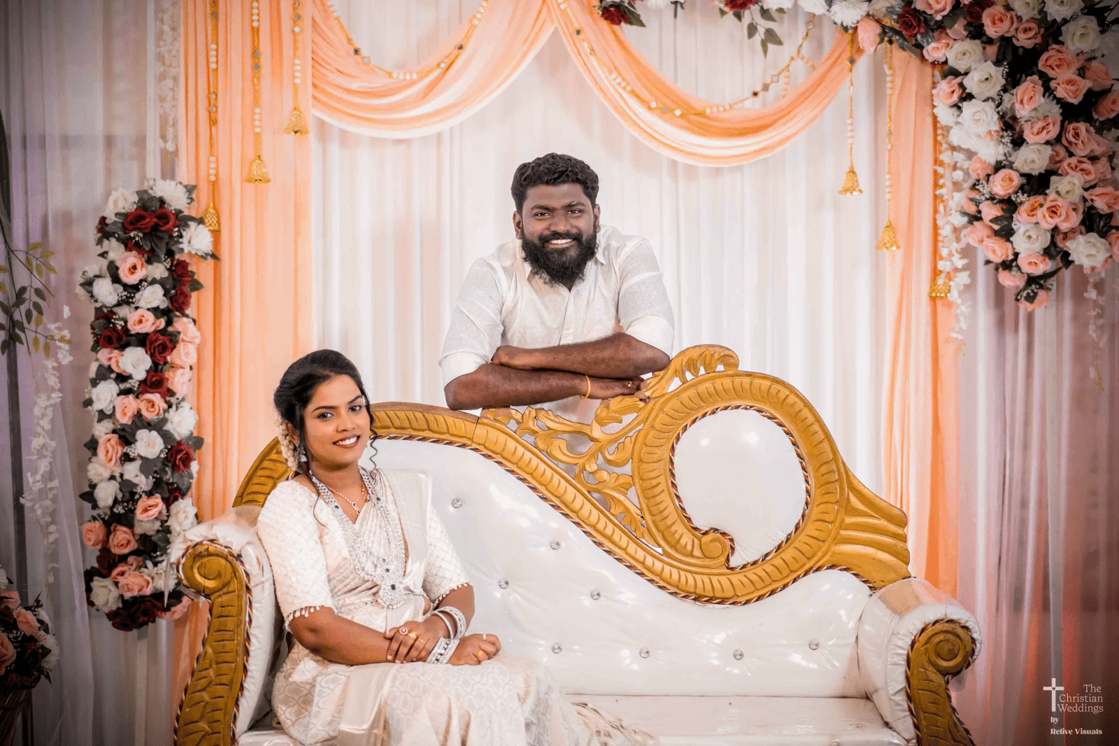 The bride sits elegantly in a chair, her wedding gown or saree gracefully draped around her. The groom stands behind her, his hands gently resting on her shoulders, both sharing a quiet moment of love.