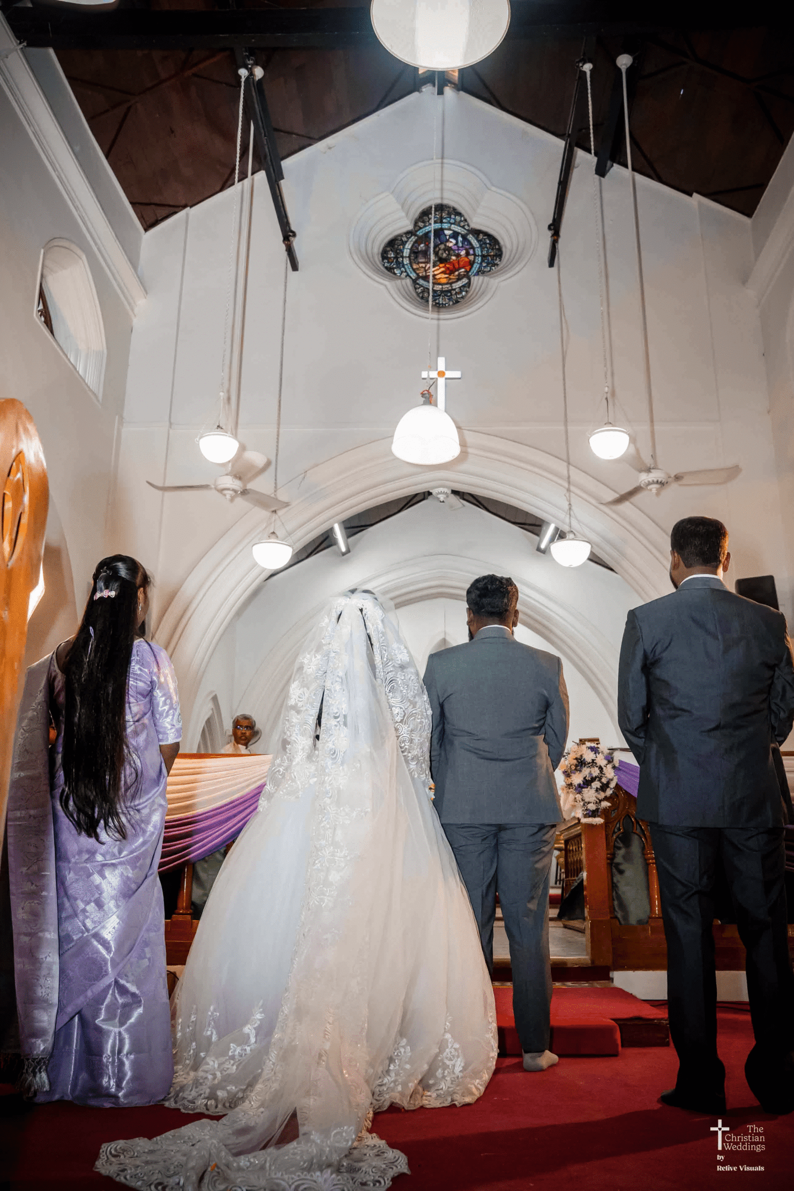 A Christian bride and groom kneel side by side, receiving blessings from the priest inside a beautifully decorated church.