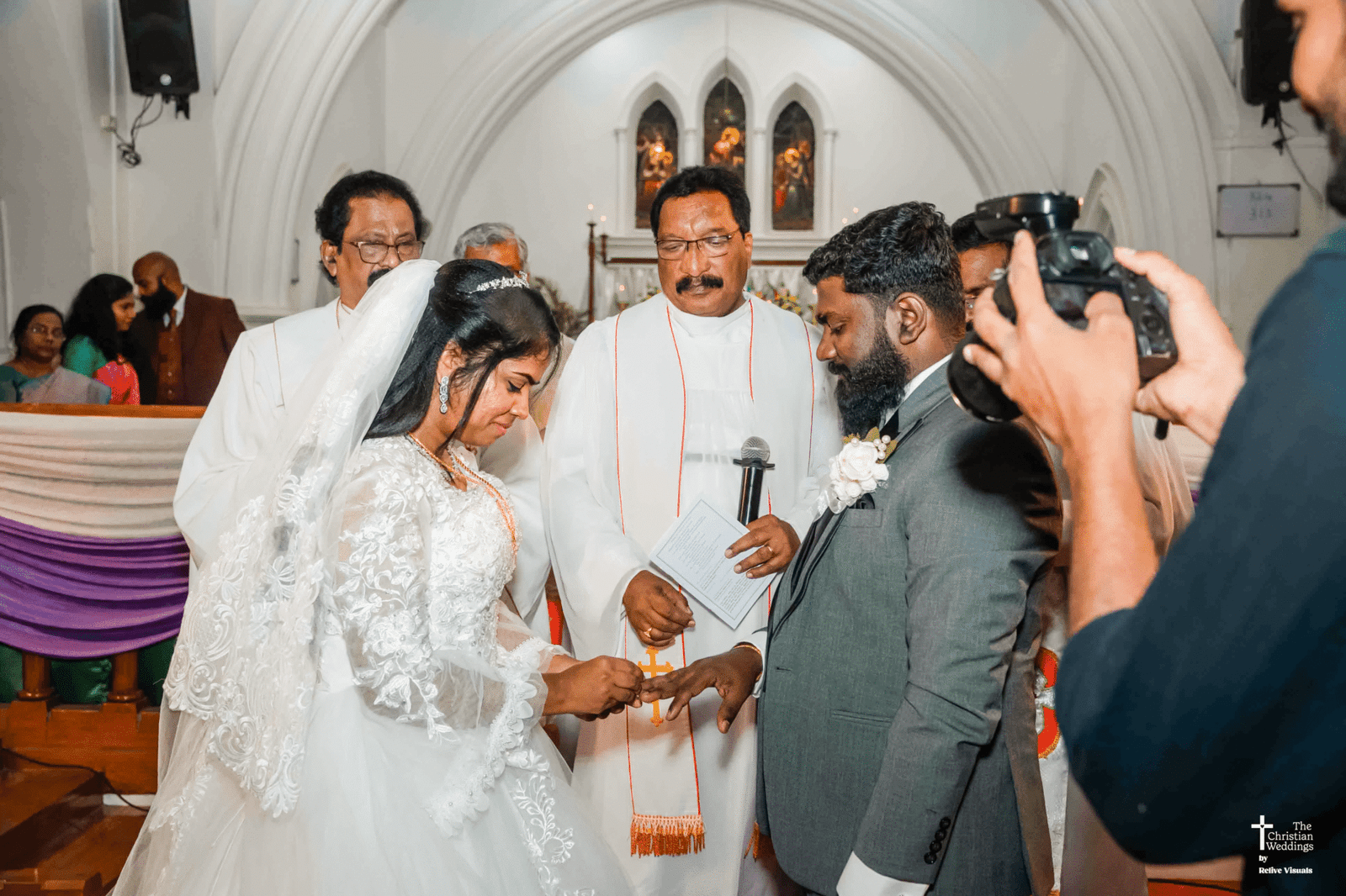 A bride and groom stand opposite each other before a statue of Jesus in a church, exchanging rings during their wedding ceremony.