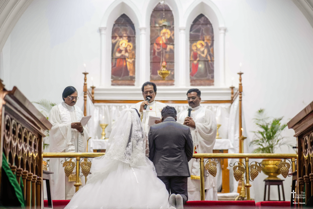 The bride and groom kneel side by side at the altar, heads bowed as the church father blesses them during their wedding ceremony.