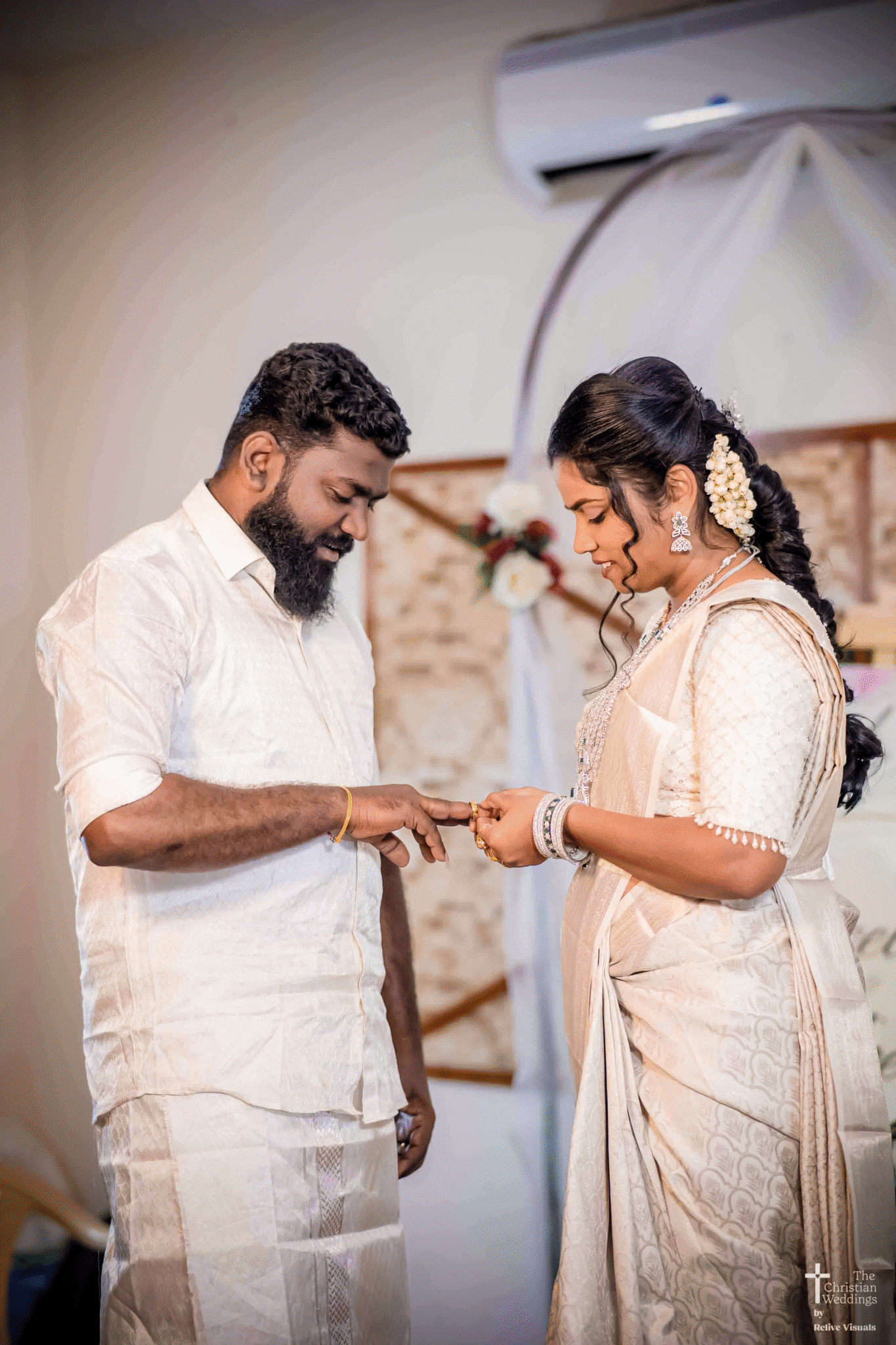 A bride and groom stand opposite each other during their engagement ceremony, as the bride places the ring on the groom’s finger with a soft, joyful smile.