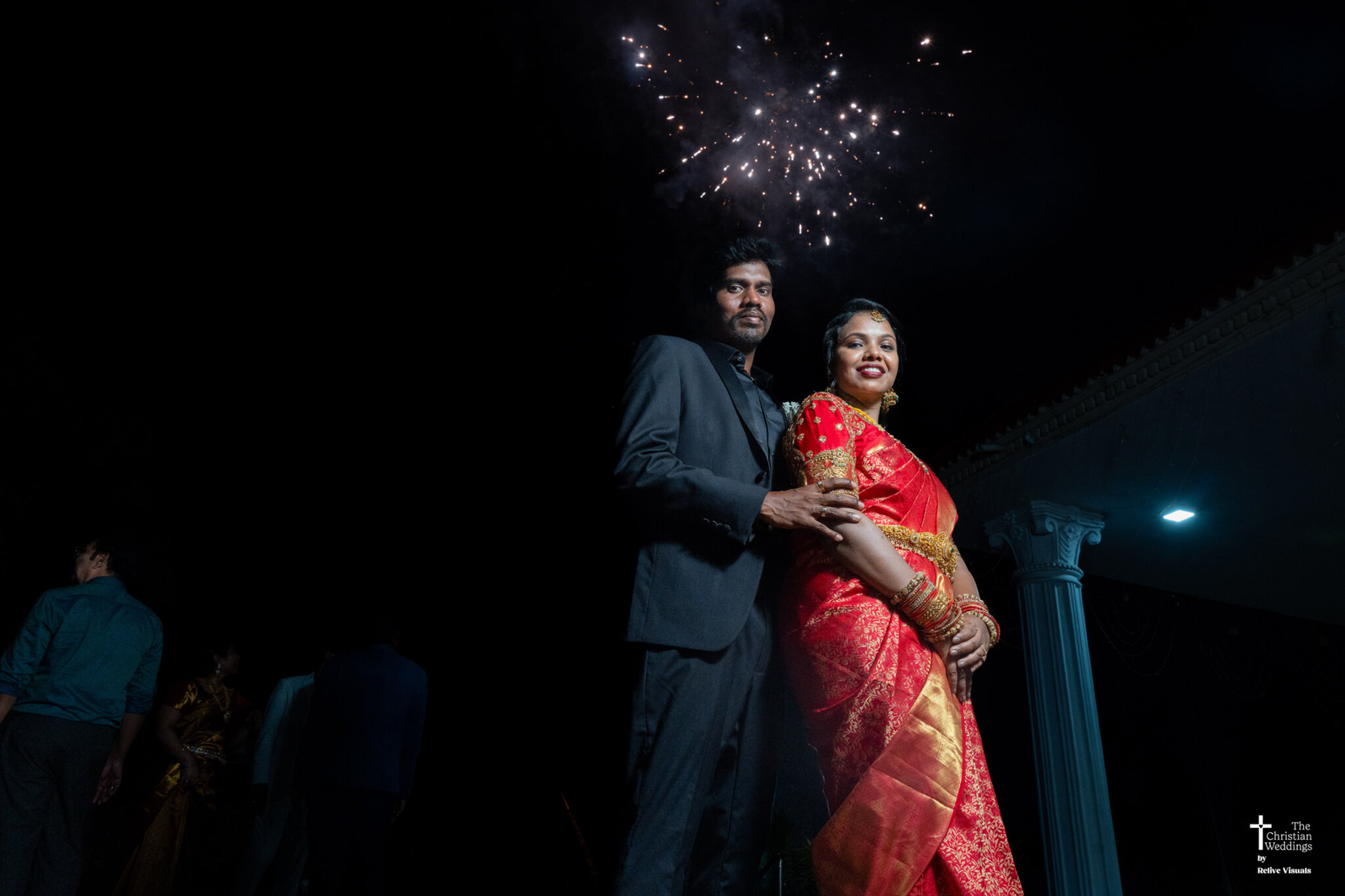Christian bride and groom standing together with colorful fireworks/crackers bursting in the night sky behind them.