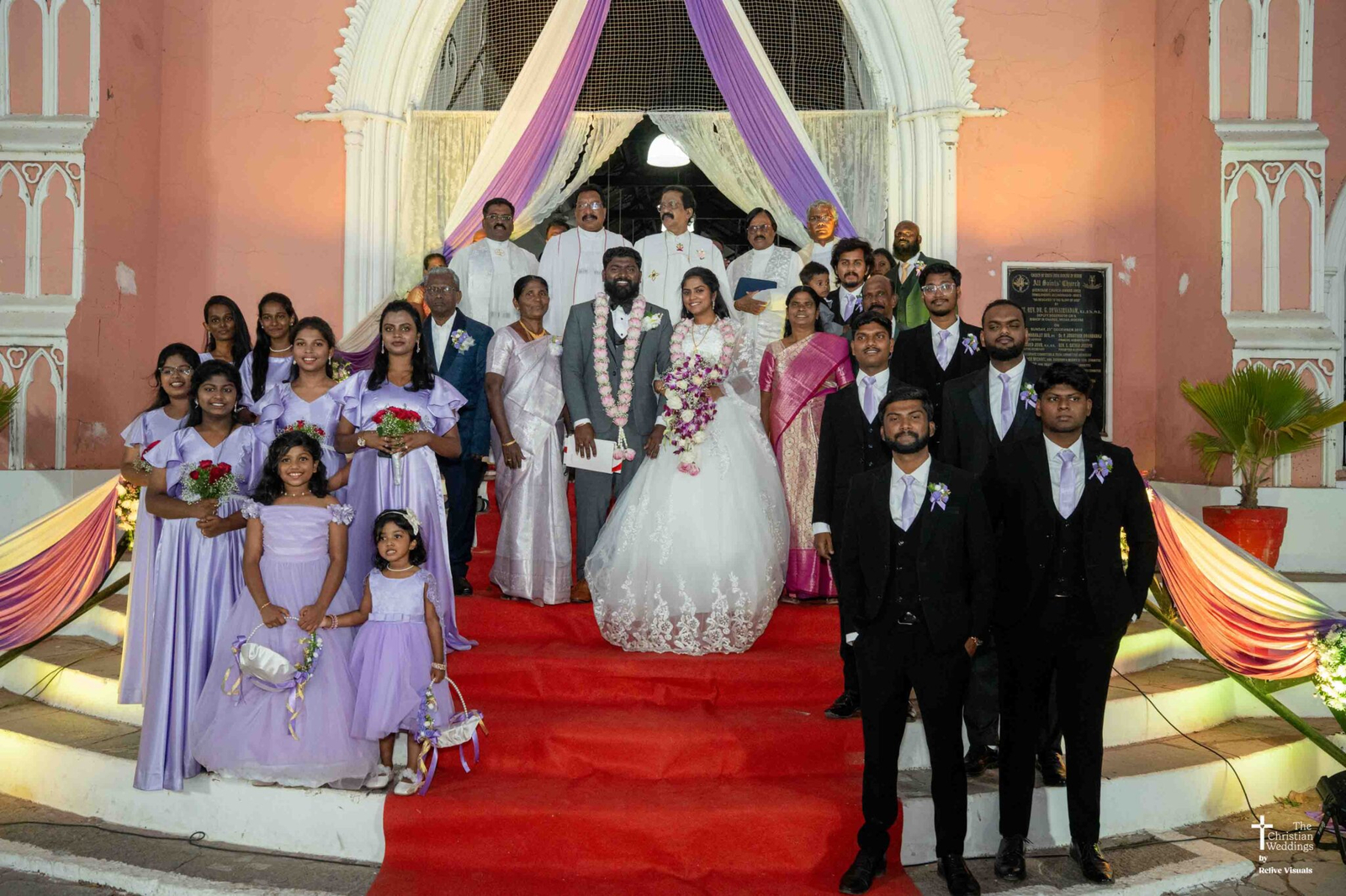 The bride and groom stand at the center, flanked by bridesmaids on both sides, all smiling joyfully and dressed in coordinated attire.