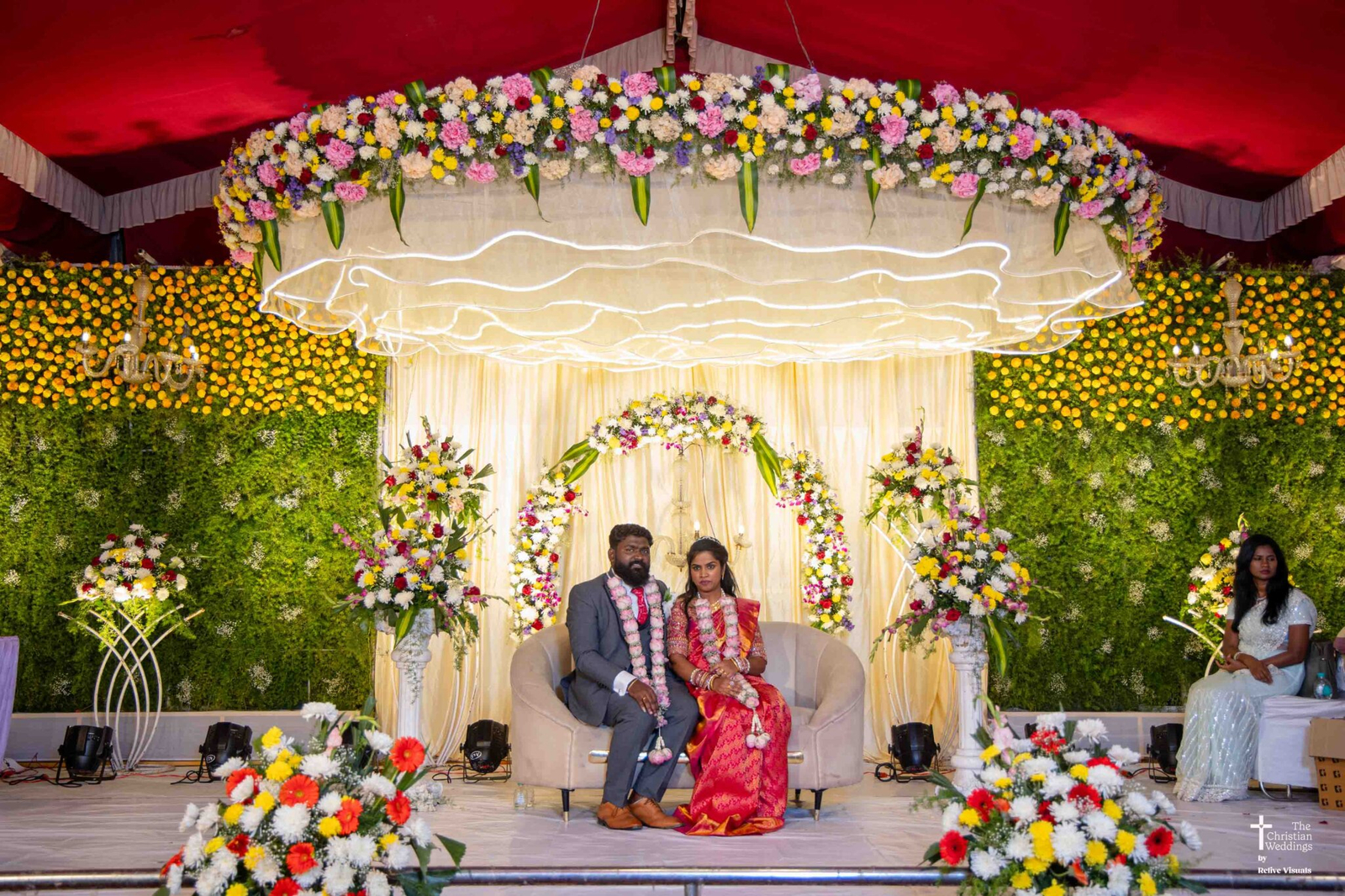 A bride and groom seated on decorated chairs in the center of a wedding stage, smiling and posing gracefully in traditional attire.